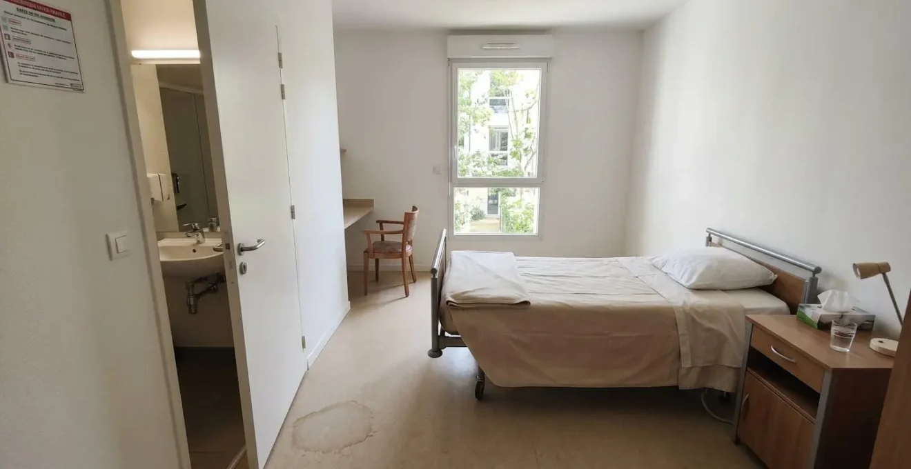 Chambre individuelle en EHPAD avec lit médicalisé, table de nuit en bois, murs blancs, porte de salle de bain entrouverte, petite trace d'humidité discrète au sol, lumière naturelle abondante, intérieur français contemporain