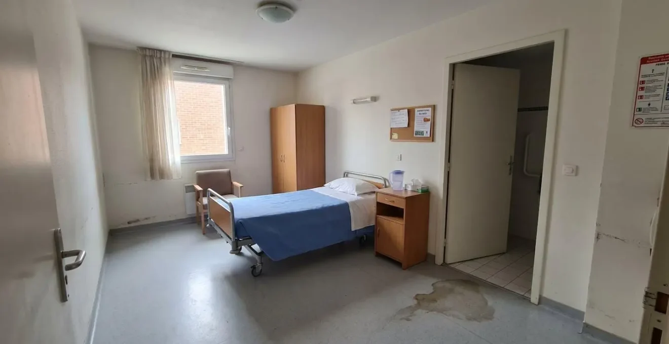 Chambre individuelle en EHPAD avec lit médicalisé, table de nuit en bois, murs blancs, porte de salle de bain entrouverte, petite trace d'humidité discrète au sol, lumière naturelle abondante, intérieur français contemporain